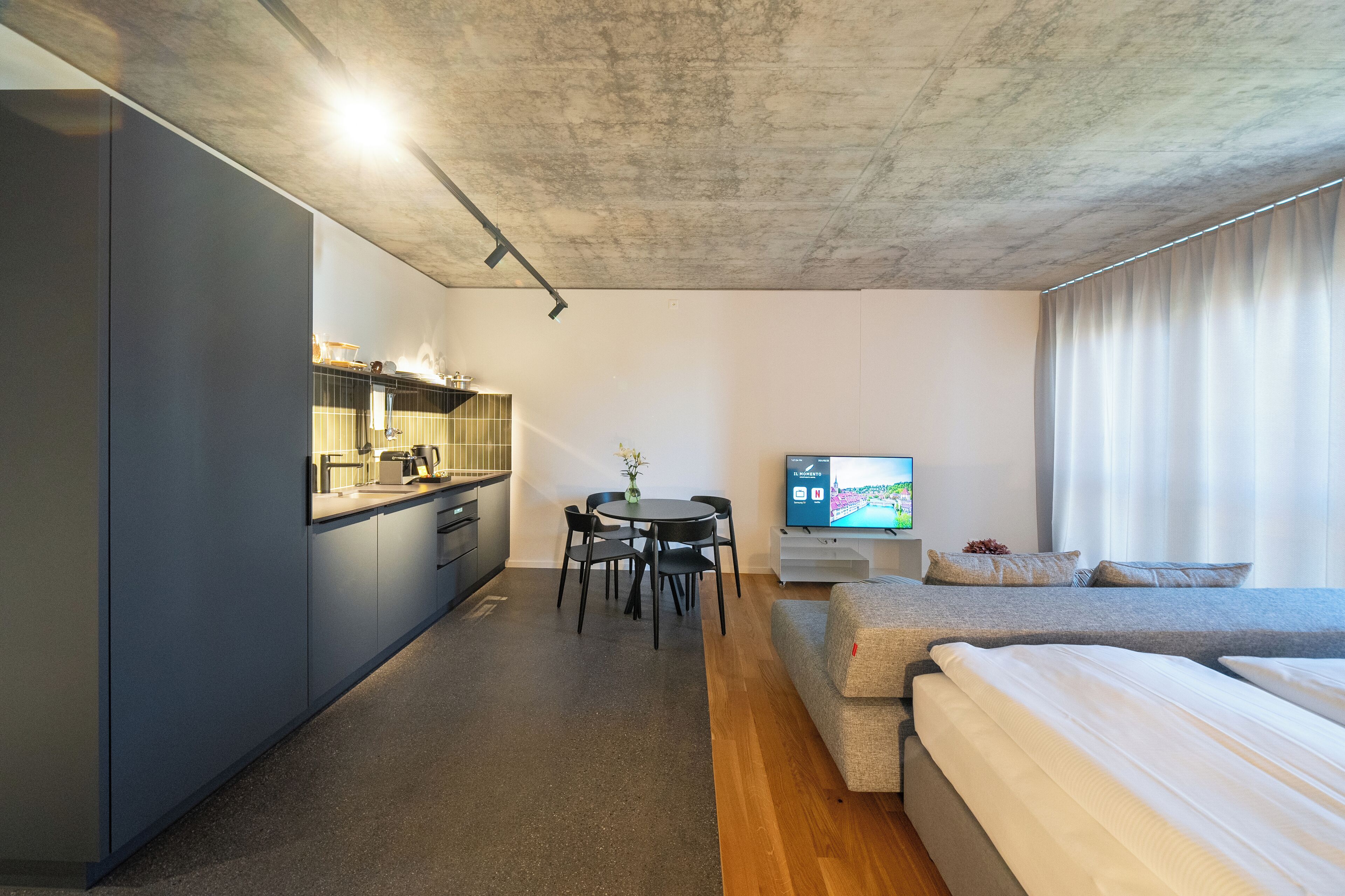 A modern hotel room with a kitchenette, a dining area, a TV, and a bed.