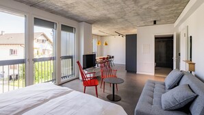 Una habitación de hotel moderna con una cama grande, un sofá gris, sillas rojas, una mesa pequeña y un balcón con vistas a casas.