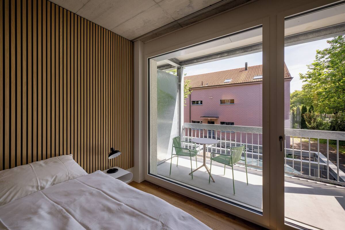 Un dormitorio con un ventanal grande, listones de madera en la pared, una cama, una mesita de noche y vistas a un balcón con sillas y un edificio al fondo.