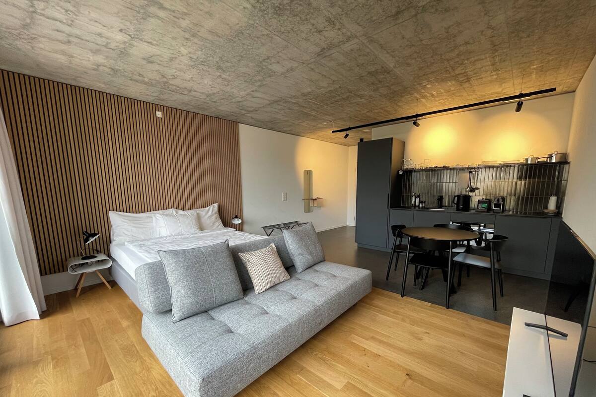 Un dormitorio moderno con una cama, un sofá gris, una mesa de comedor y una zona de cocina.