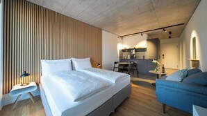 Una habitación de hotel moderna con una cama grande, un sofá azul, una pequeña mesa de comedor y una kitchenette al fondo.
