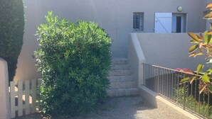 A courtyard with a gravel path, a white picket fence, a green hedge, and a staircase leading to a building with a white door and window. - 2 ROOM APARTMENT, SEMAPHORE, SAINTE MAXIME (Sainte-Maxime)