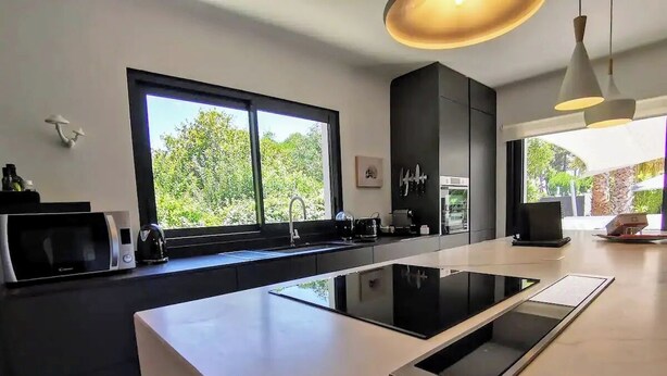 A modern kitchen with a large island, black cabinets, and a window providing a view of greenery. - 4 Bedroom Villa in Anglet (Anglet)