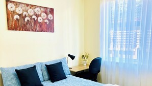 A neatly made bed with a blue floral bedspread, a white quilt, and two folded towels. There is a desk with a lamp and a potted plant, and a window with sheer curtains.