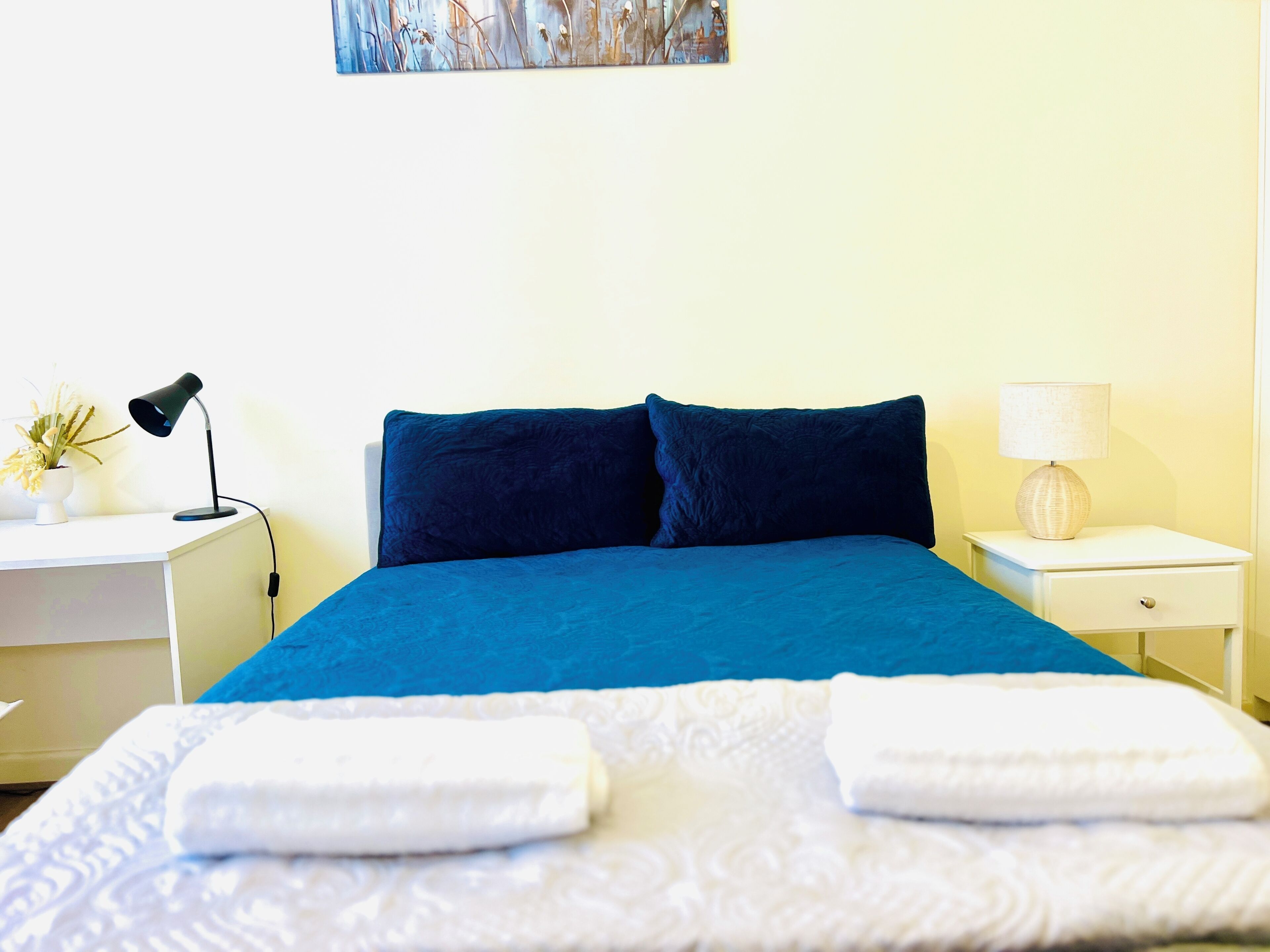 Cama bem arrumada com colcha azul e duas almofadas, mesa de cabeceira branca com abajur, uma pequena planta em vaso e um quadro na parede.