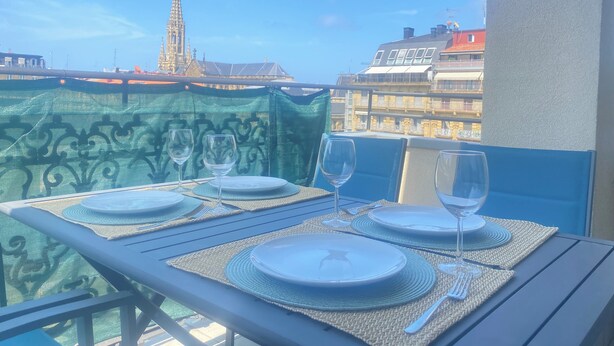 A balcony table set with plates, glasses, and cutlery, overlooking a cityscape with a prominent tower. - Mikain Cathedral View (Donostia)