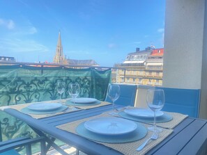 Une table de balcon, dressée avec des assiettes, des verres et des couverts, d’où l’on peut admirer un panorama urbain dominé par une tour imposante.