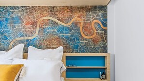 Een hotelkamer met een grote kaart aan de muur, een bed met witte beddengoed, een mosterdkleurig kussen, en een blauwe plank met spullen erop.