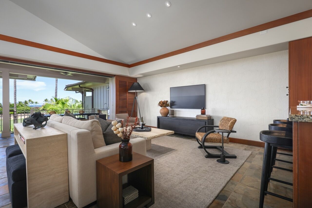 Una sala moderna con sofá, mesa de centro y televisor. Hay un balcón con vistas a palmeras.