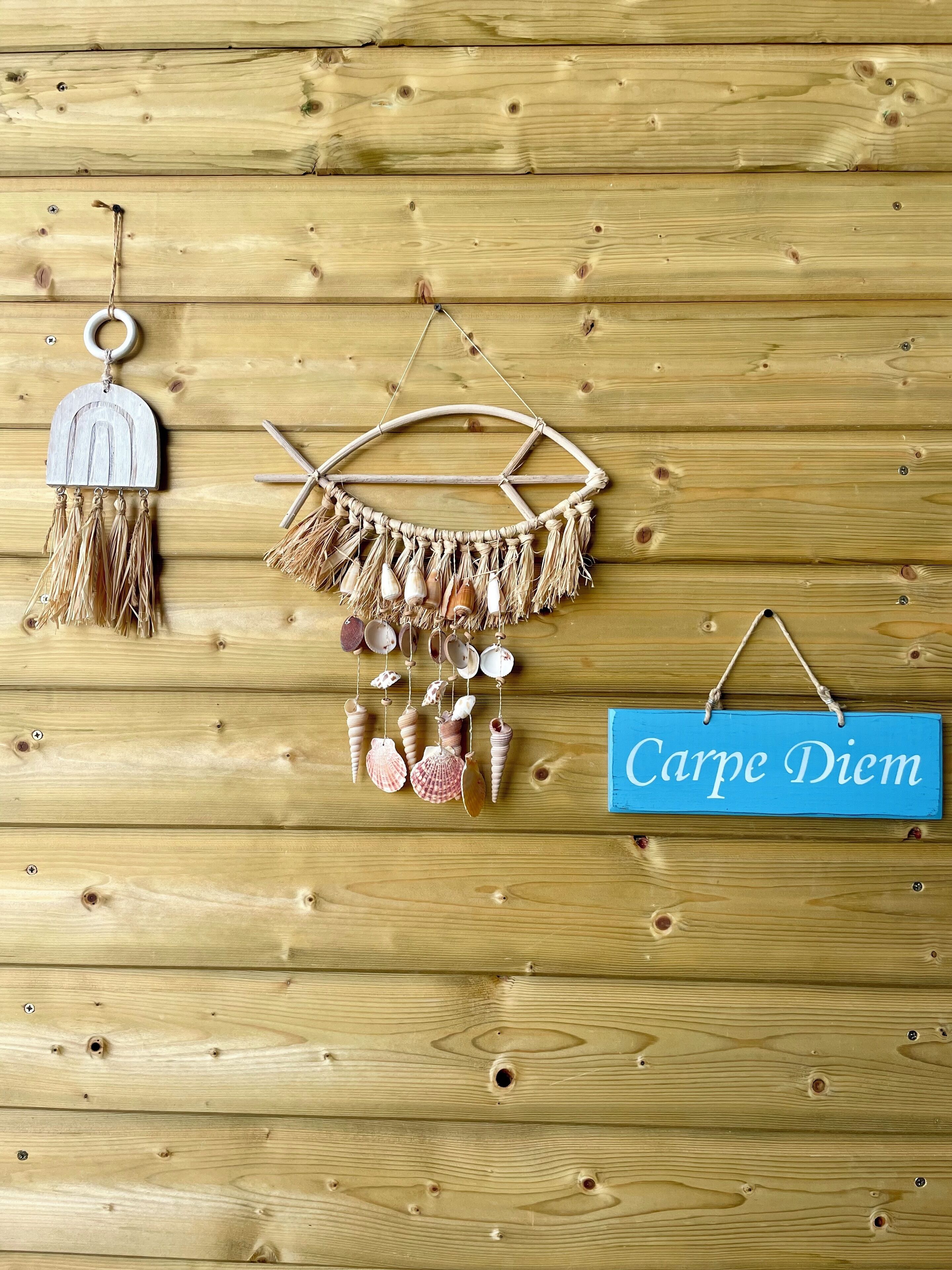 Una parete in legno decorata con tre elementi: un arazzo in macramè, un arazzo in macramè con conchiglie e un cartello blu con testo.