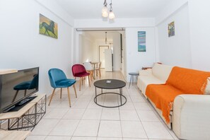 Un salon moderne avec un téléviseur à écran plat, une table basse et un canapé agrémenté d’un plaid orange. On y trouve deux fauteuils, un bleu et un rouge, ainsi qu’une table à manger à l’arrière-plan.