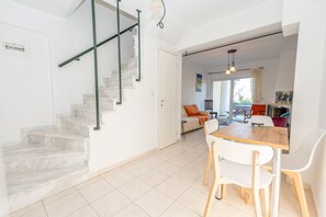 Un intérieur moderne avec un escalier en marbre, un coin repas comprenant une table en bois et des chaises blanches, ainsi qu’un canapé agrémenté de coussins orange.