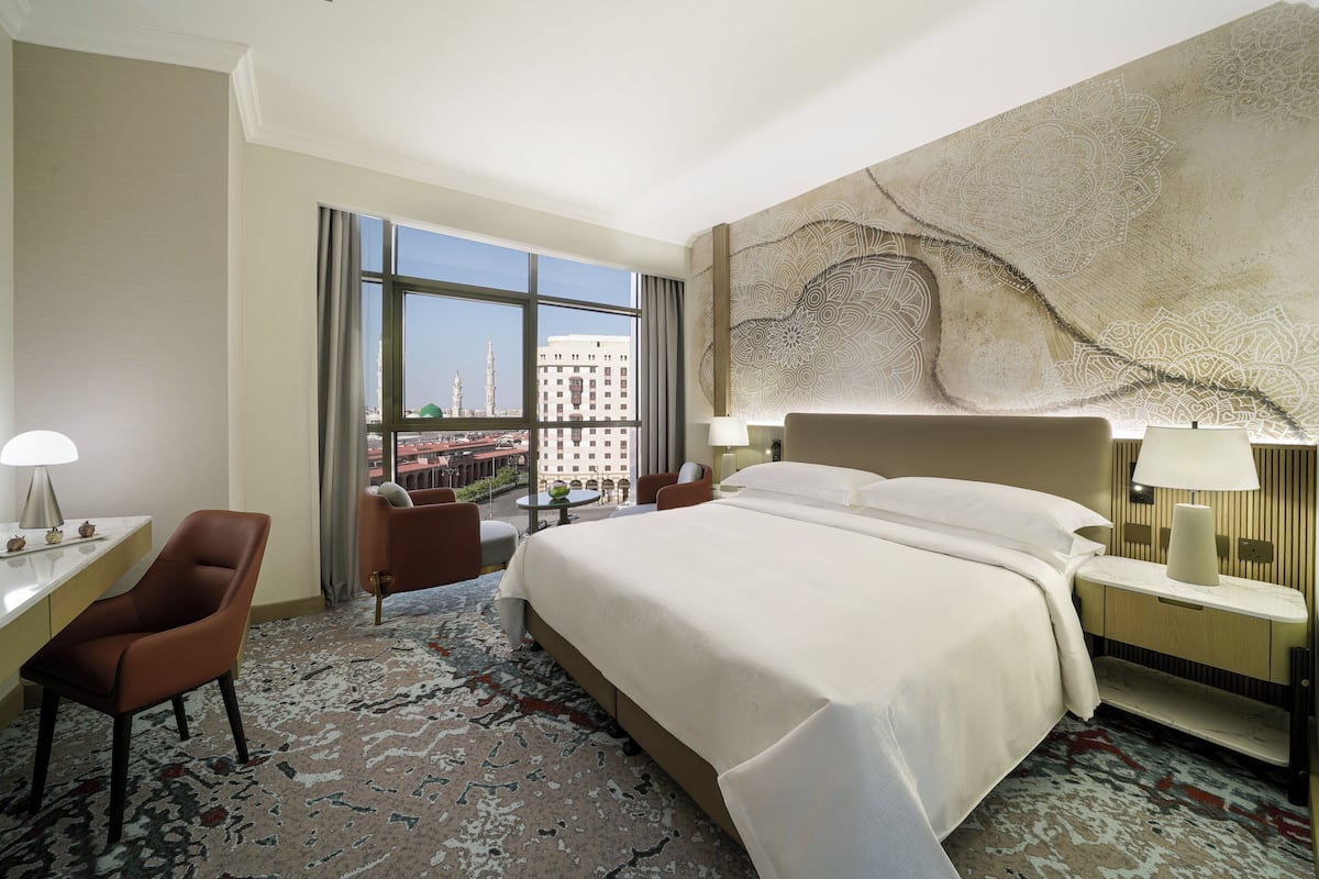 A hotel room with a large bed, a desk, a chair, and a view of a cityscape through large windows.