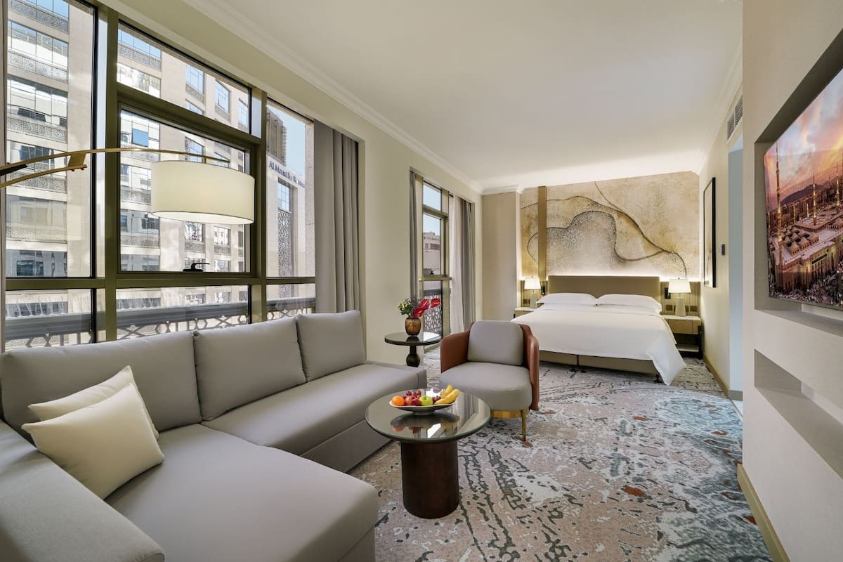 A hotel room with a sofa, armchair, bed, and a painting of a cityscape.