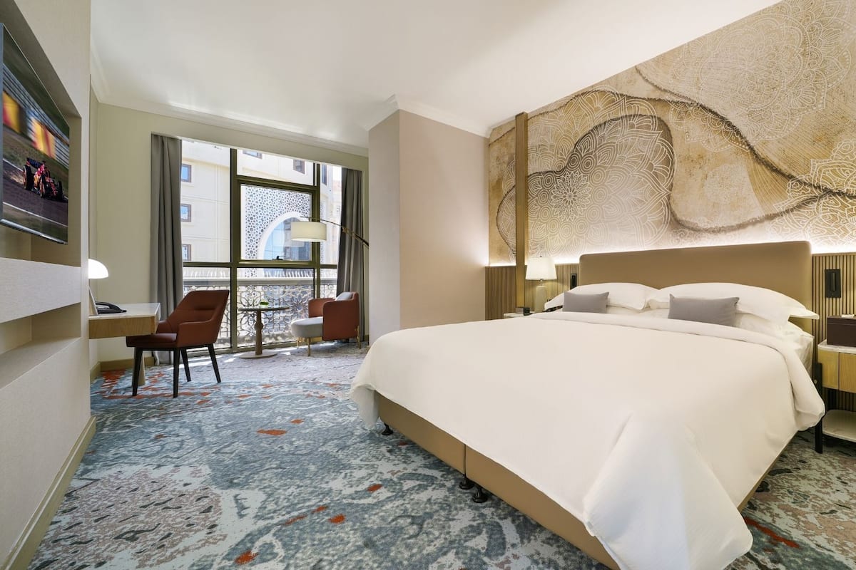 A hotel room with a large bed, a desk with a chair, a nightstand, and a wall with a decorative pattern.