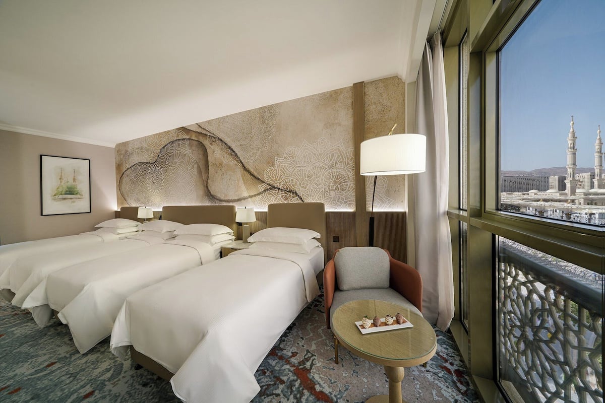 A hotel room with two beds, a chair, a small table with snacks, and a large window with a view of a cityscape.