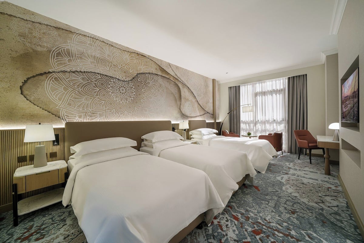 A hotel room with three beds, a large decorative wall, a desk with a lamp, and a chair.