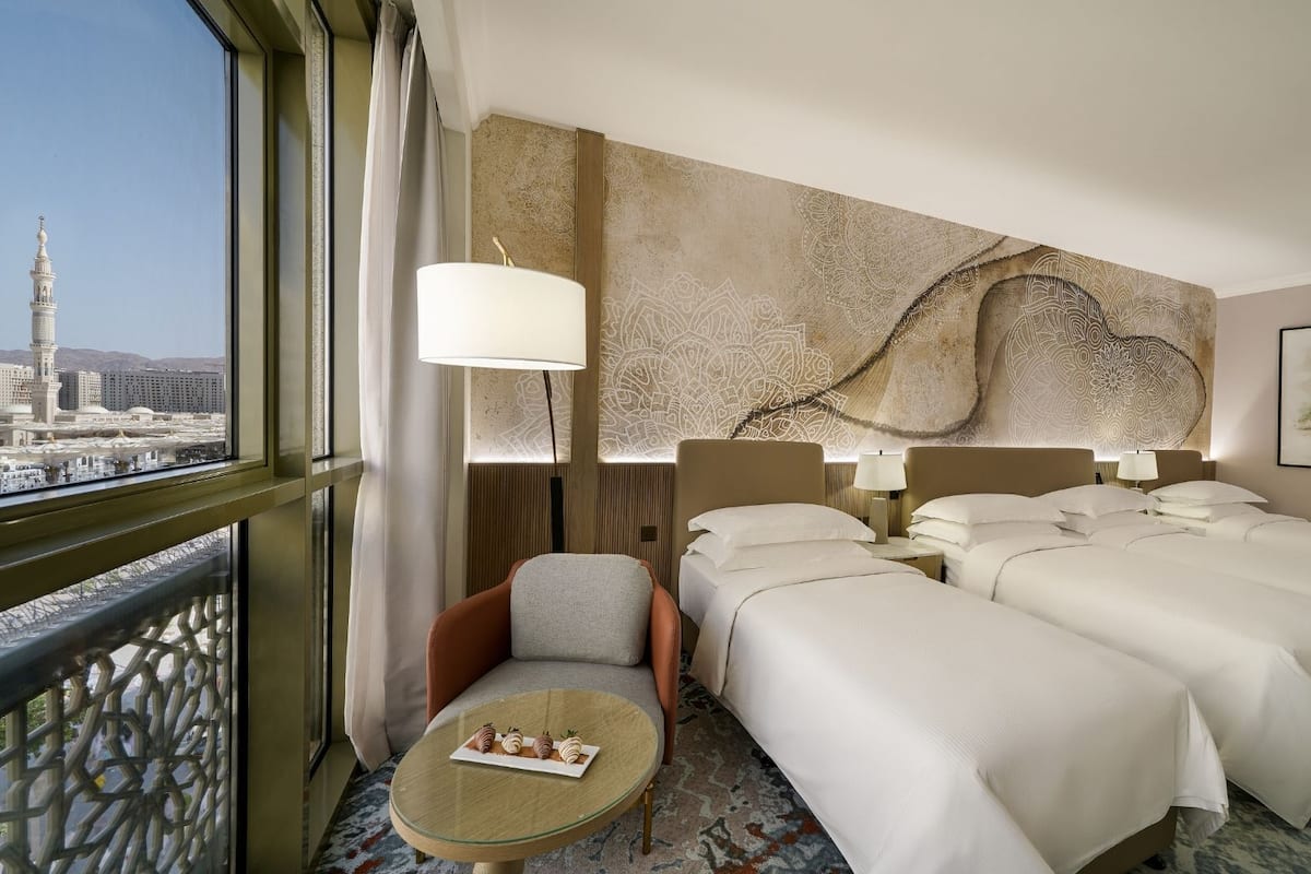 A hotel room with two beds, a sofa, a small table with a tray of snacks, a large window with a view of a mosque, and a decorative wall.