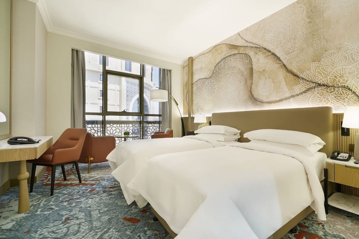 A hotel room with a large bed, a desk with a telephone, a chair, and a view of the city through large windows.