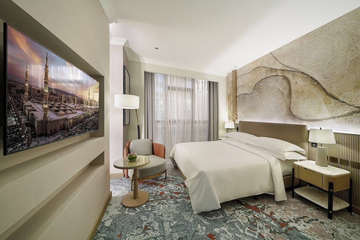 A hotel room with a large bed, a chair, a small table, and a wall-mounted TV displaying a cityscape.