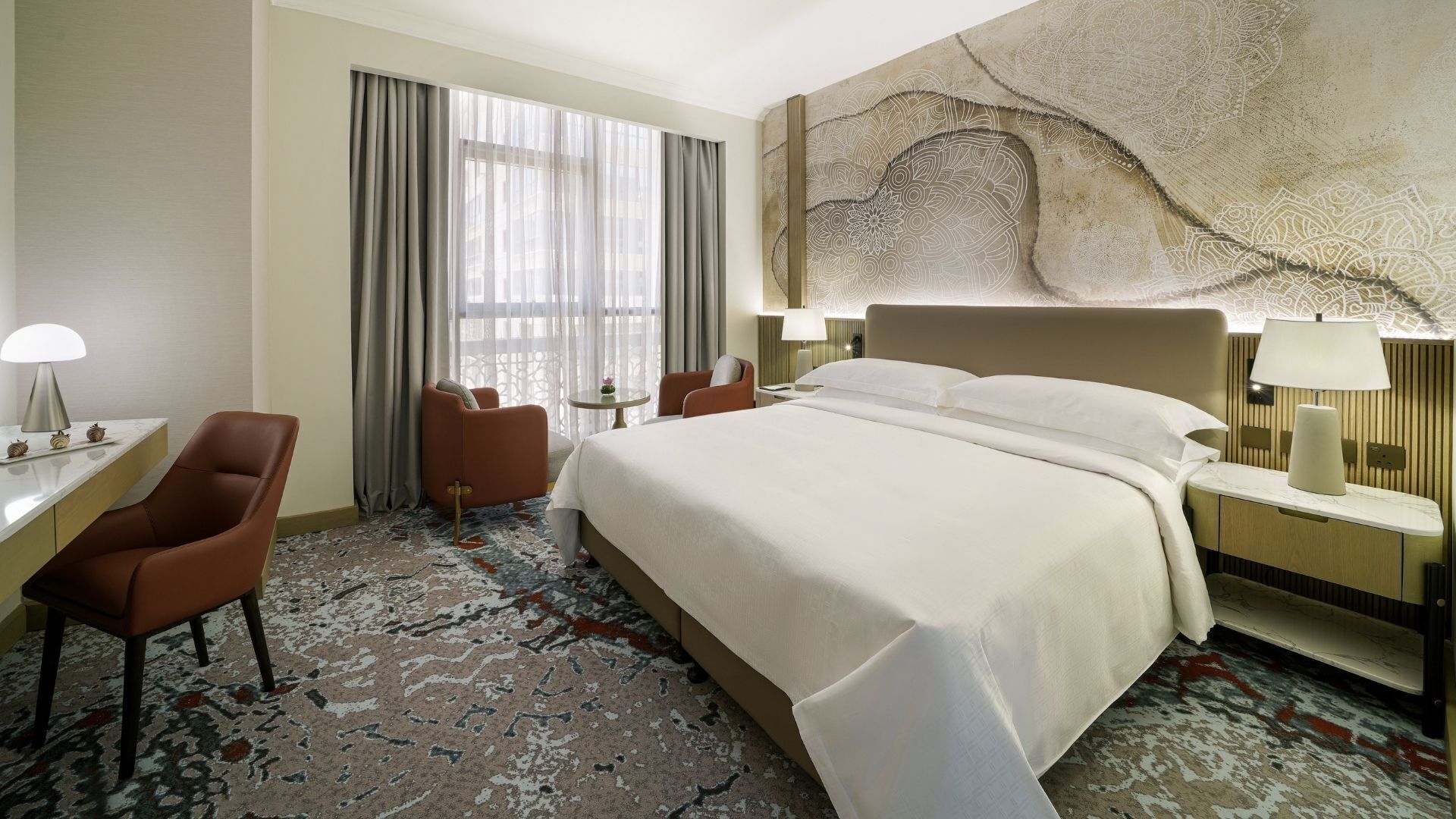 A hotel room with a large bed, a desk with a lamp, two chairs, and a large abstract wall art piece.