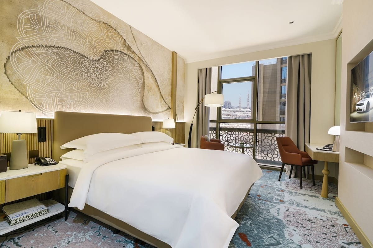 A hotel room with a large bed, a desk with a chair, a TV, and a view of the city through large windows.