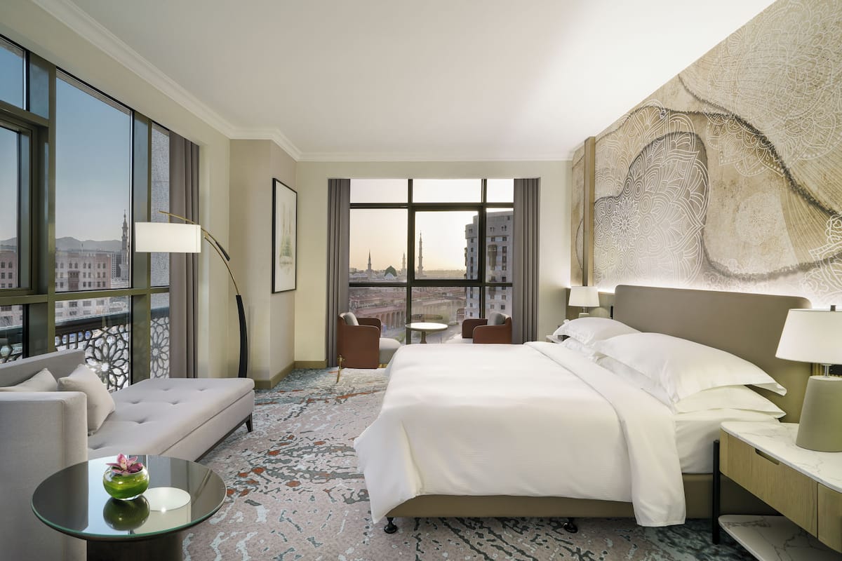 A hotel room with a large bed, a sofa, a small table, and a view of a cityscape through large windows.