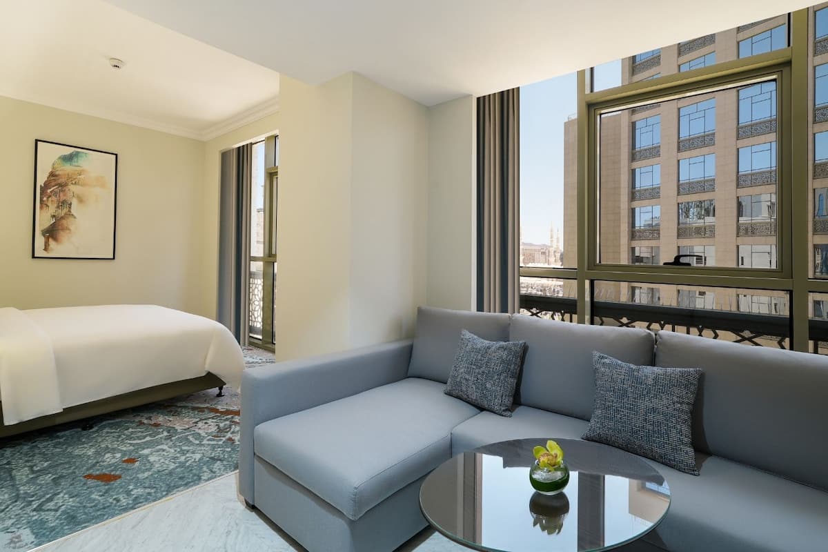 A hotel room with a bed, a sofa, a small table, and a large window offering a city view.