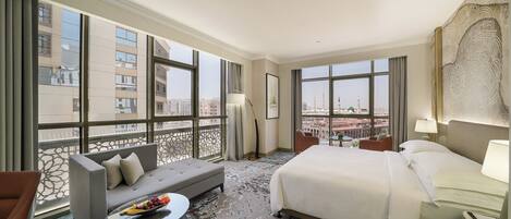 A hotel room with a large bed, a sofa, a small table with fruit, and a view of the cityscape through large windows.