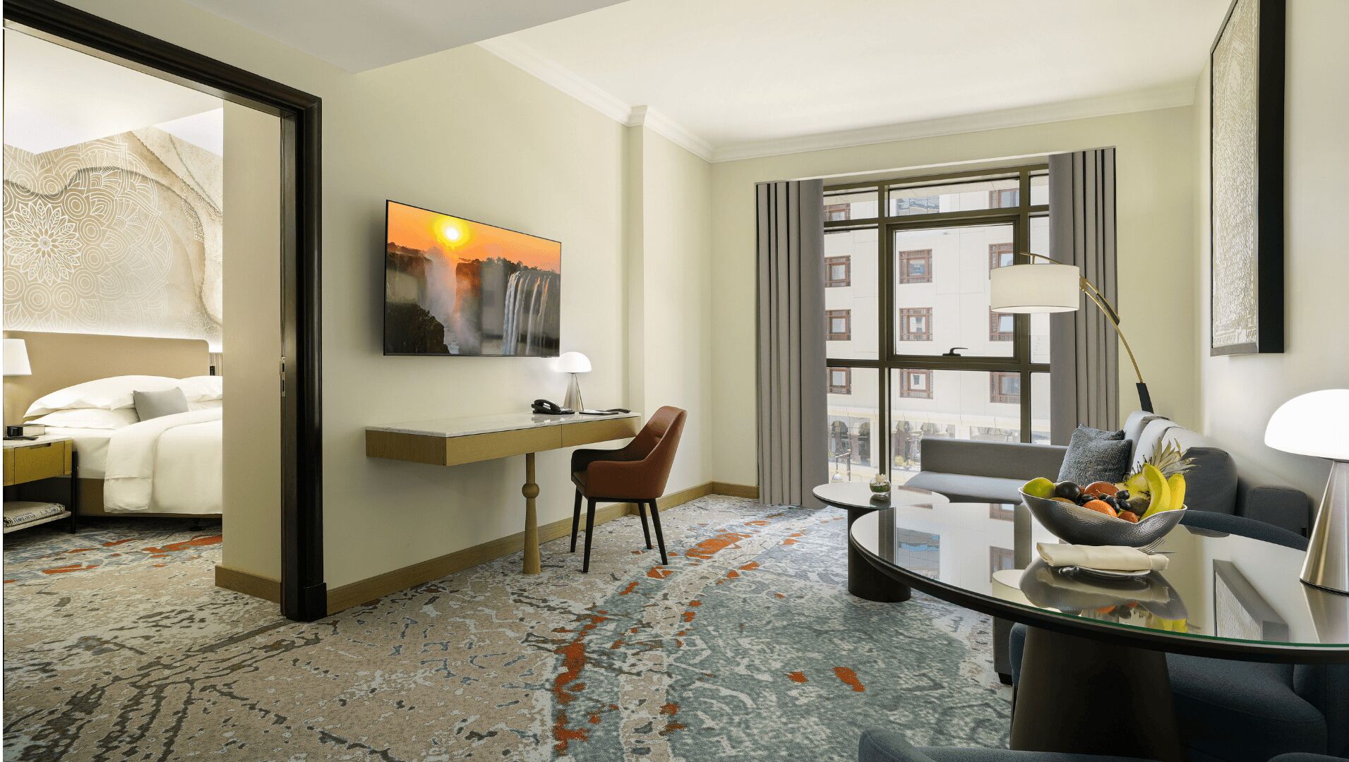 Habitación de hotel con cama, escritorio, silla, mesa de comedor con frutero, un cuadro y un ventanal con vistas a edificios.