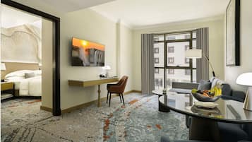 Habitación de hotel con cama, escritorio, silla, mesa de comedor con frutero, un cuadro y un ventanal con vistas a edificios.