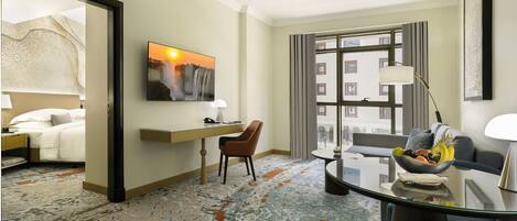 Habitación de hotel con cama, escritorio, silla, mesa de comedor con frutero, un cuadro y un ventanal con vistas a edificios.