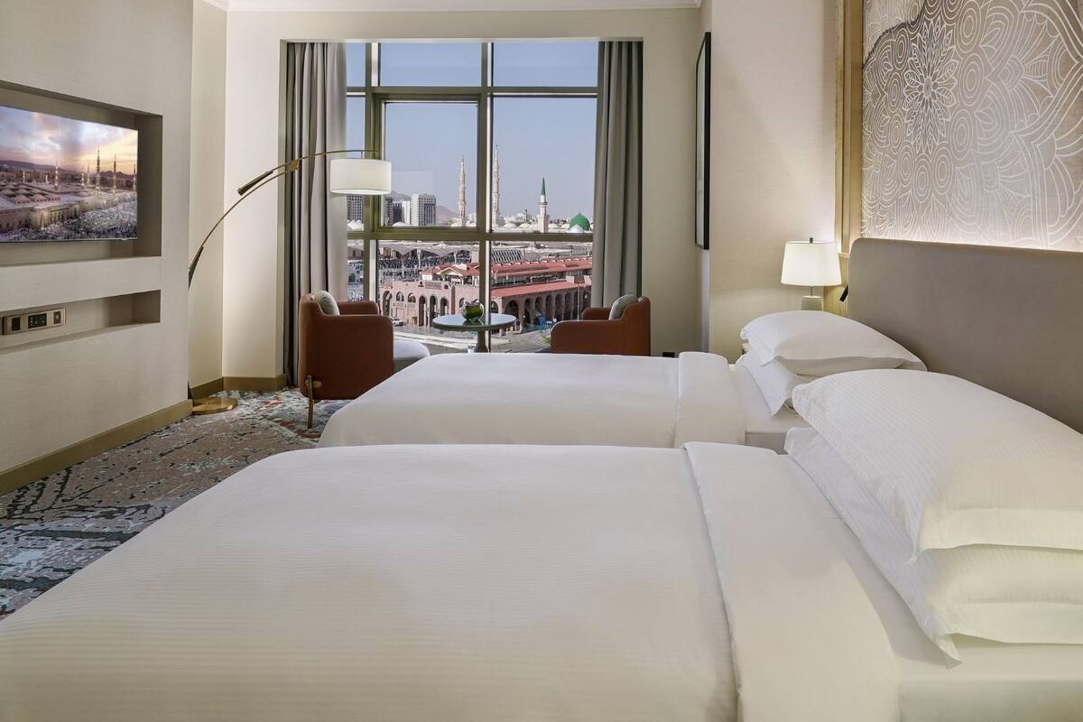 A hotel room with two beds, a large window with a city view, a flat-screen TV on the wall, and a patterned wall hanging.