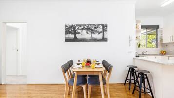 Ein Essbereich mit einem für vier Personen gedeckten Tisch, einem Schwarz-Weiß-Baumgemälde an der Wand und einer durchgehenden Tür erkennbaren Küche.