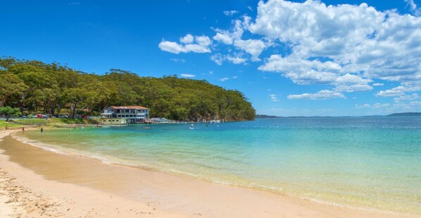 A beach with clear turquoise water, a sandy shore, and a building with a balcony overlooking the sea.