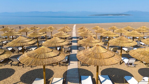 Una spiaggia con file di lettini e ombrelloni di paglia, una passerella in legno e vista sul mare e sulle montagne in lontananza.