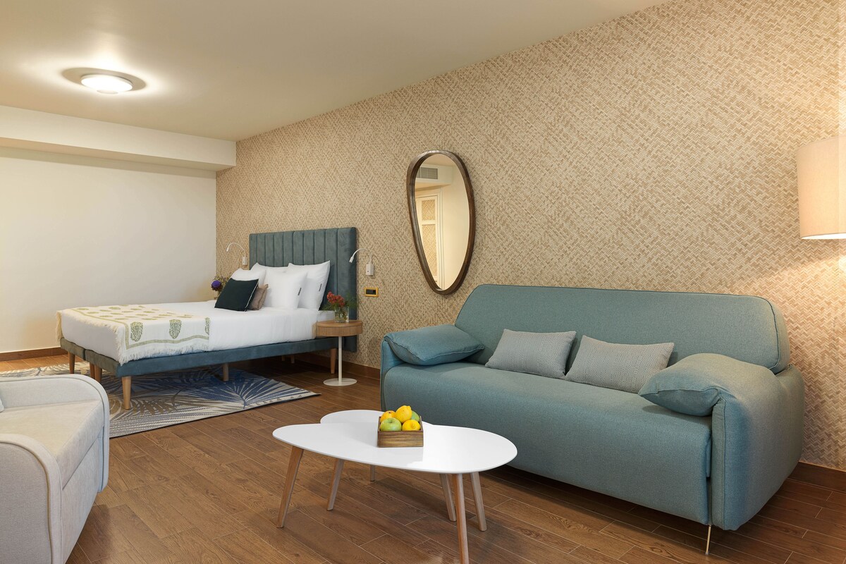 A hotel room with a bed, a sofa, a small table with fruit, and a mirror on the wall.