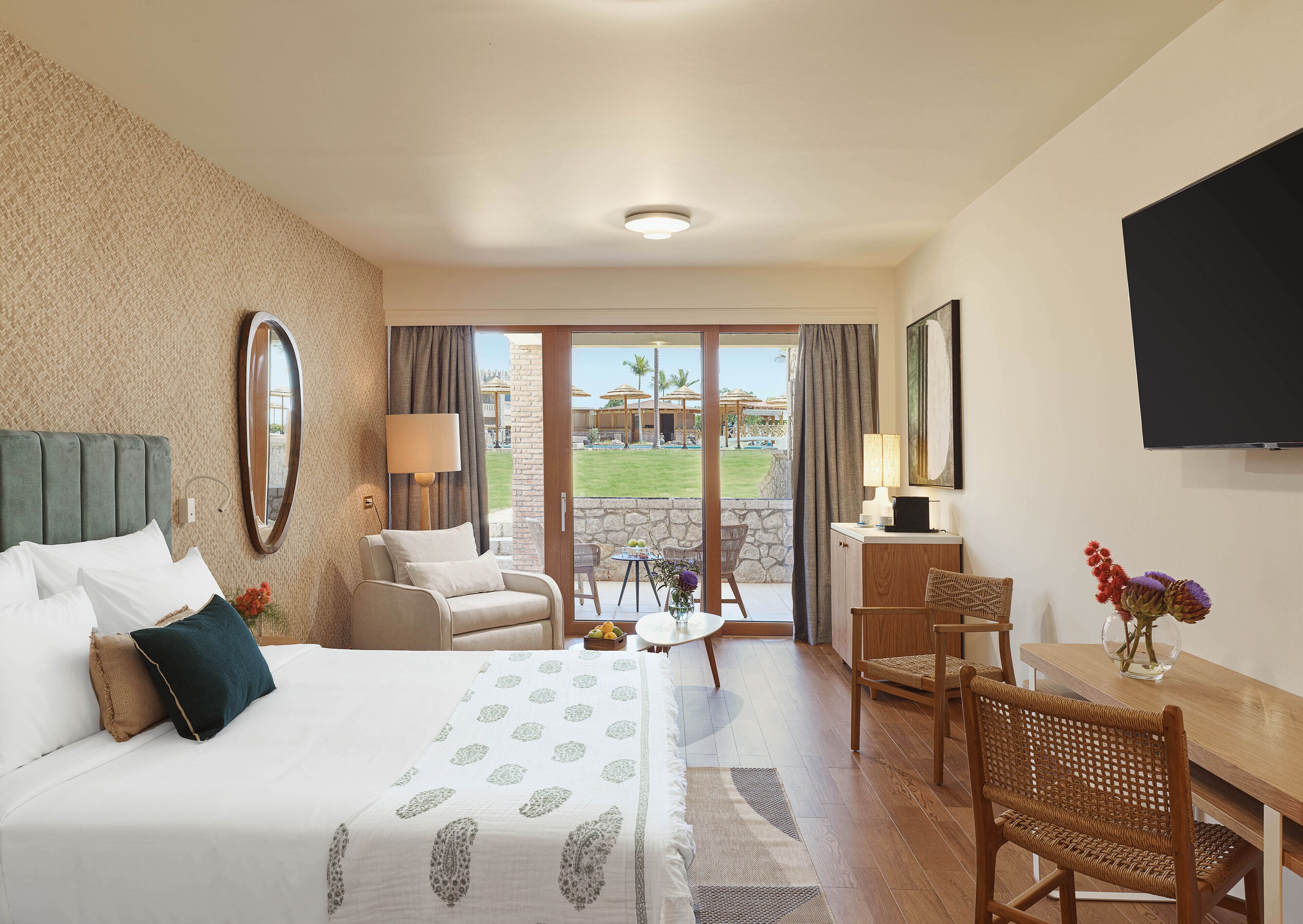 A hotel room with a bed, a desk, a chair, a TV, and a view of the outdoor area.