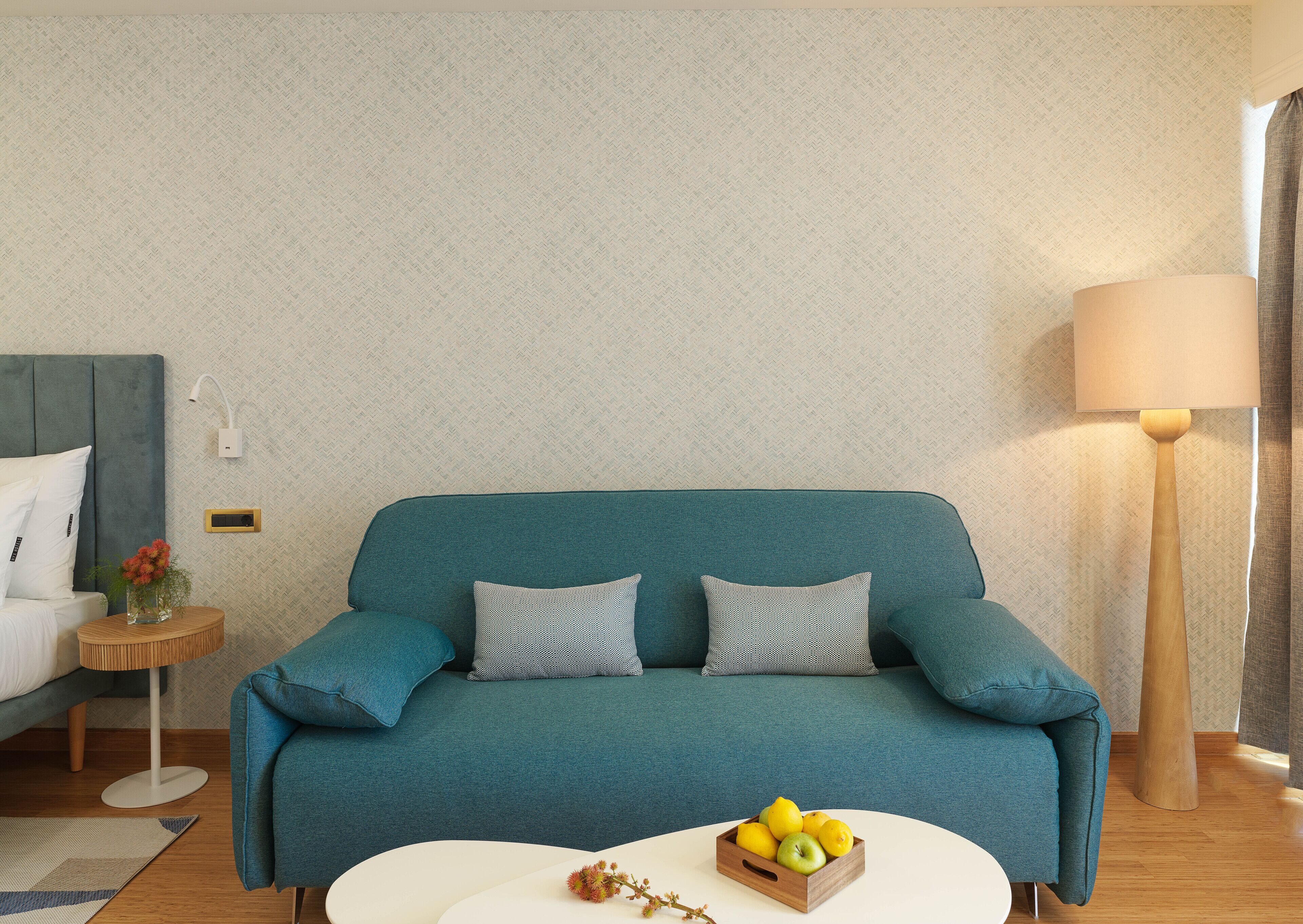 Ein smaragdgrünes Sofa mit gemusterten Kissen, ein kleiner Holztisch mit einer Blumenvase und ein weißer Couchtisch mit Obstschale.