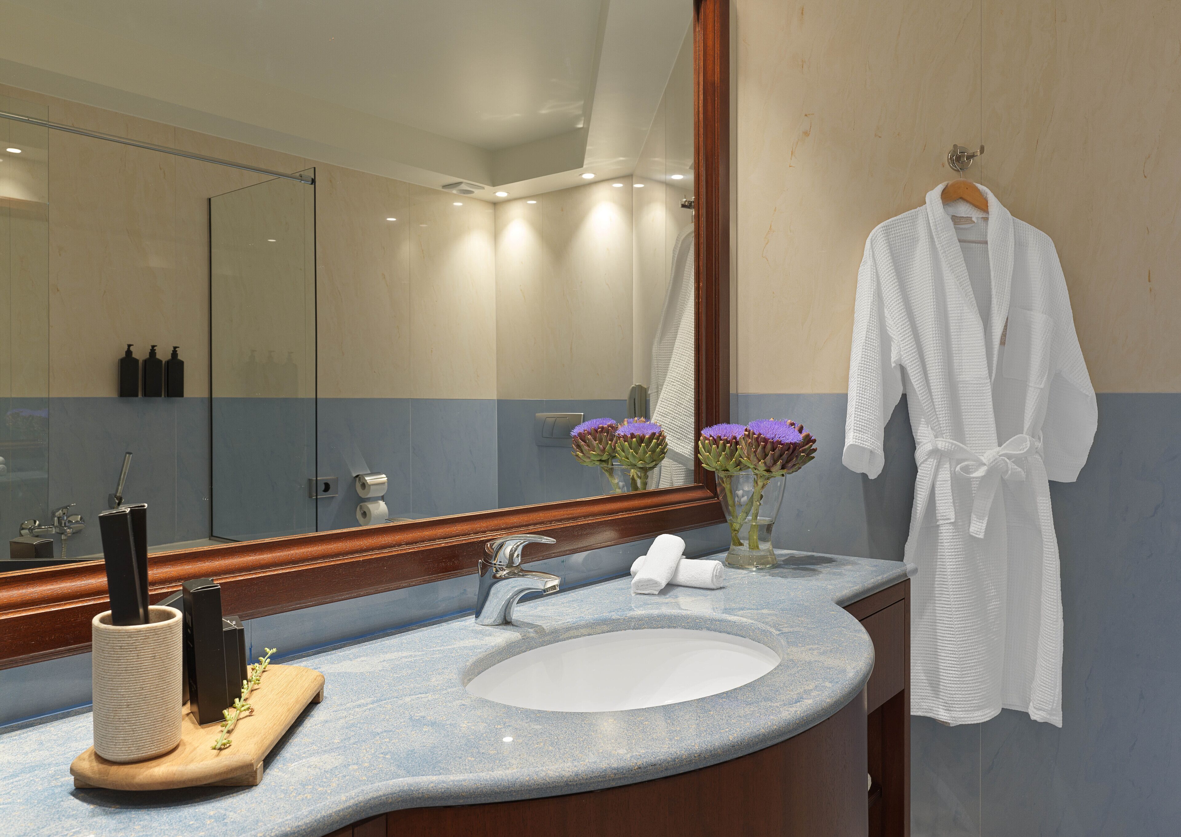 Une salle de bain avec un grand miroir, un lavabo, un plateau en bois avec une brosse, et une robe accrochée au mur.