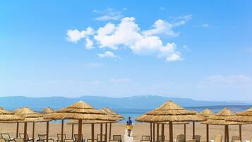 A beach with rows of sun loungers and thatched umbrellas, a clear sky, and a person walking on a path.