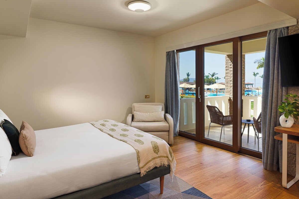 A hotel room with a bed, a chair, a TV, and a view of an outdoor pool and palm trees.