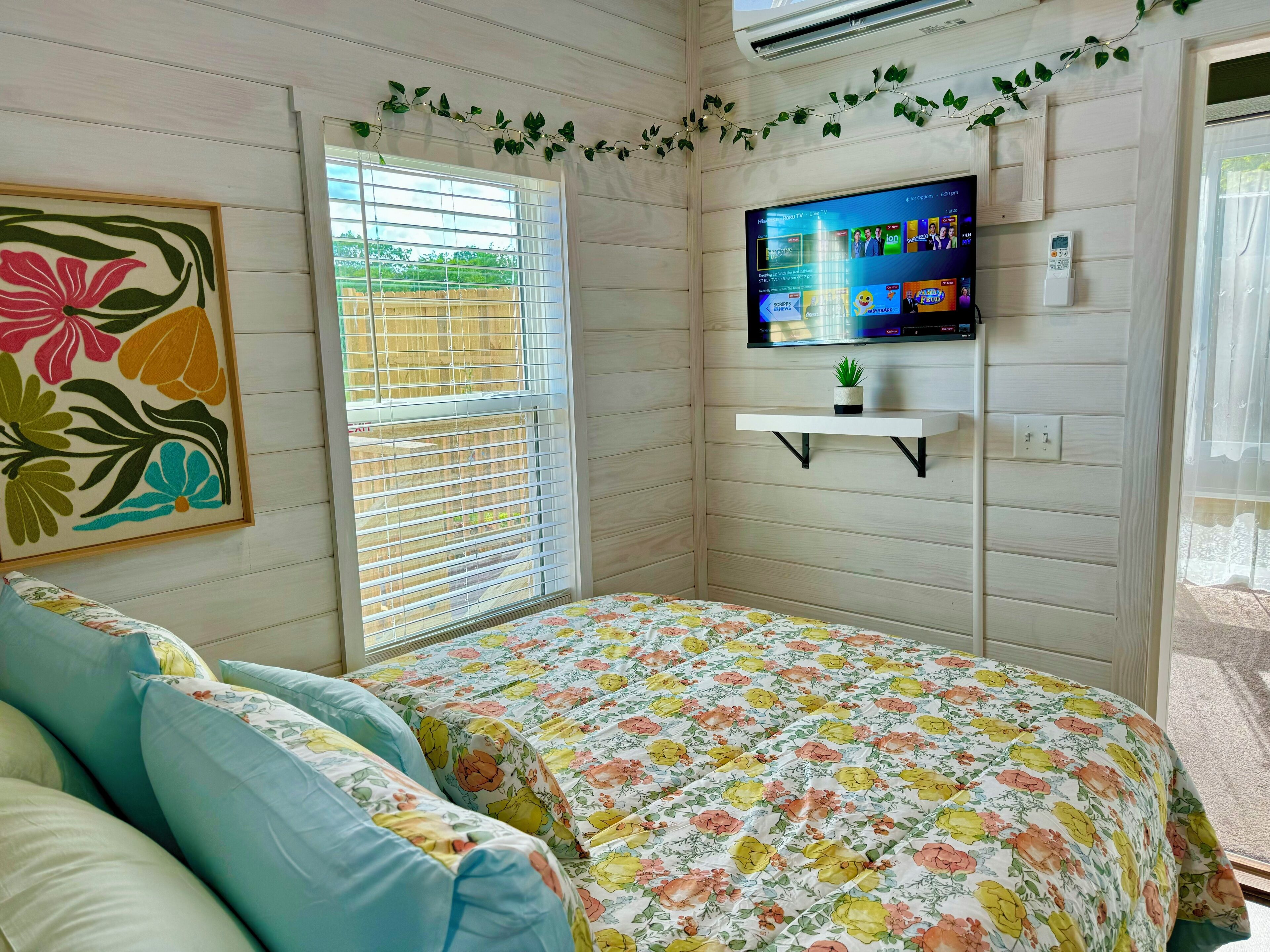 Une chambre à coucher avec un lit, un téléviseur fixé au mur, une fenêtre avec des stores et un tableau représentant des motifs floraux accroché au mur.