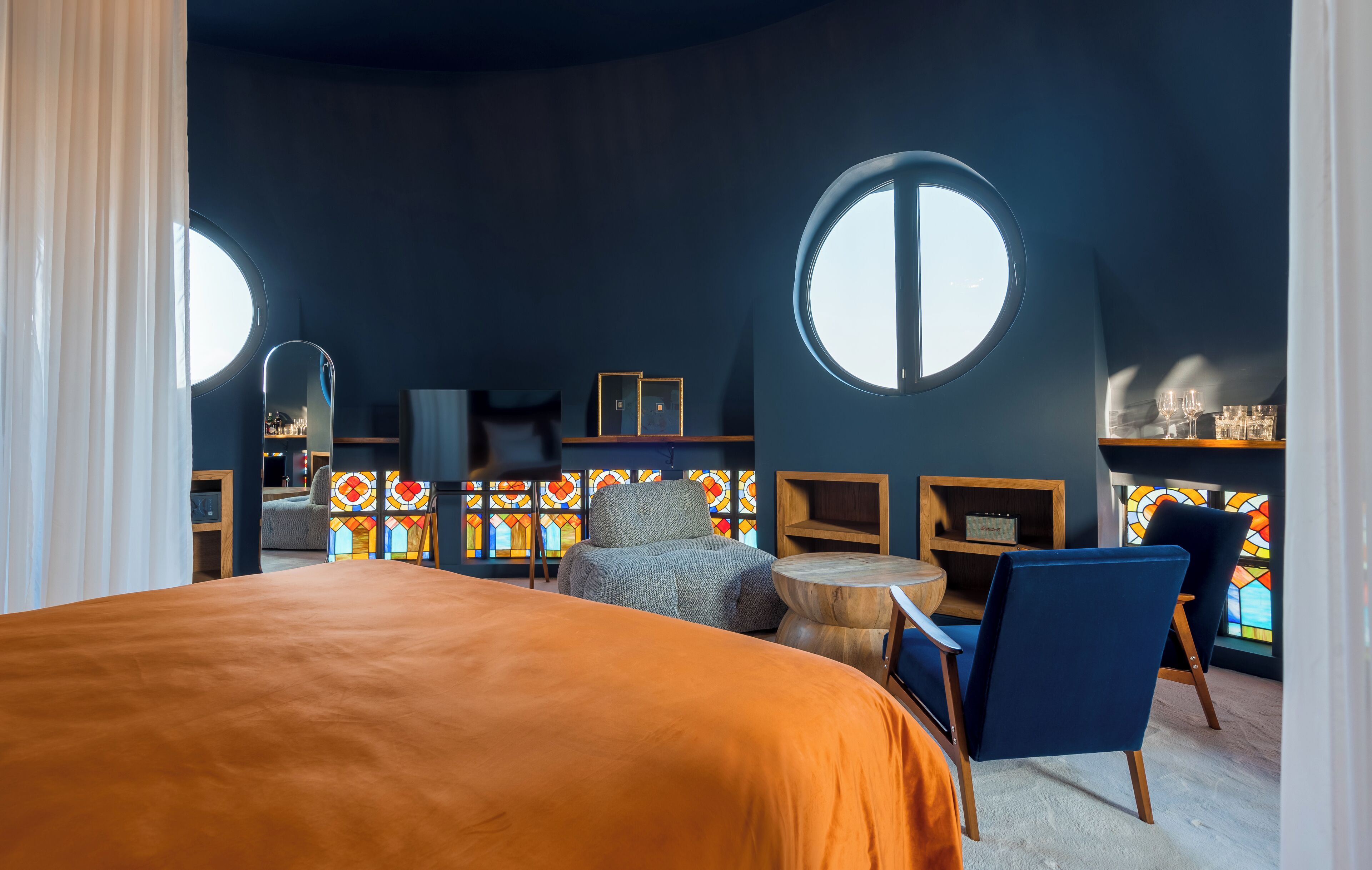 Een kamer met een groot bed, twee ronde ramen, een stoel, een kleine tafel en een muur met glas-in-loodramen.
