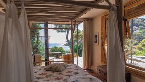 Una camera da letto con un letto, tende, una testata in legno, una finestra vista mare e un tavolo con sedie all'esterno.