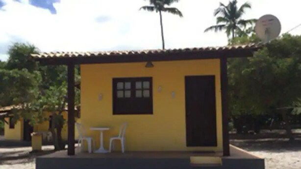 Pieni keltainen rakennus, jossa on laatoitettu katto, ovi ja ikkuna. Sen ulkopuolella on kaksi valkoista muovituolia.