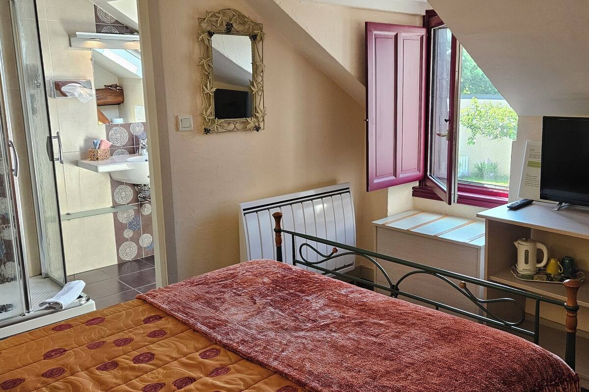 A bedroom with a bed, a TV, a desk, and a window with open shutters.