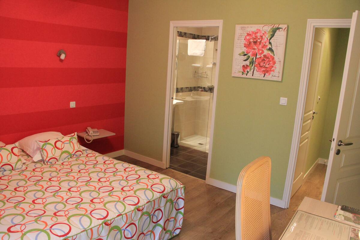 A bedroom with a bed, a nightstand, a chair, and a bathroom with a shower.
