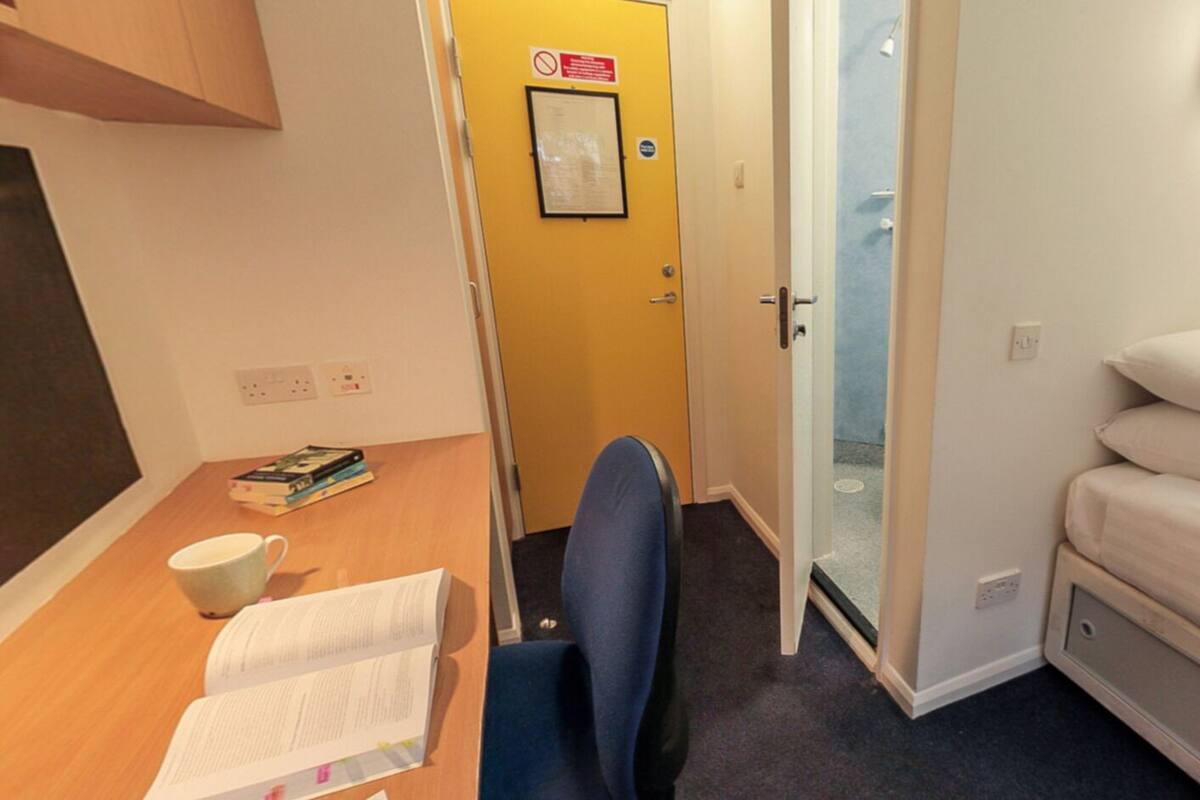 A compact room with a bed, a desk with books and a cup, and a door leading to a bathroom.