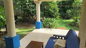 Een overdekt terras met een blauwe bank en een houten tafel, omgeven door zuilen en een betegelde vloer.